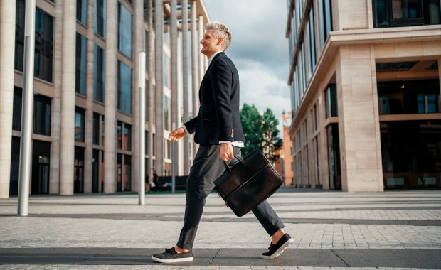 A trim, middle-aged caucasian man wearing a black blazer with gray dress pants and gray loafers with white soles walks briskly through a city plaza while smiling and carrying a briefcase.