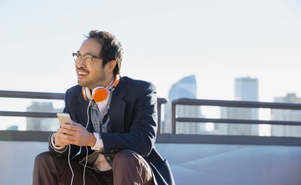 An Asian-American male in a blazer, dress shirt, and khaki slacks sits on a bench with orange-and-white headphones around his neck, holding a smartphone, and looking off in the distance smiling, with a city silhouette behind him.